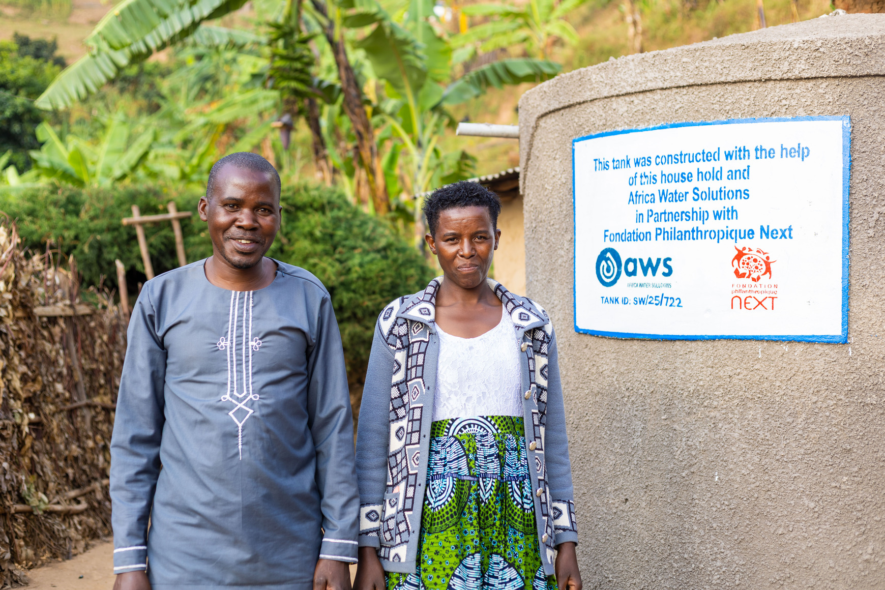 Emmanuel and his wife stand besides a rainwater harvesting tank